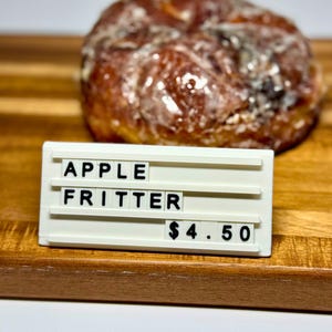 Puede incluir: Un buñuelo de manzana glaseado sobre una tabla de madera. El buñuelo es de color marrón dorado. Un letrero blanco frente al buñuelo dice "APPLE FRITTER" y "4,50 $."