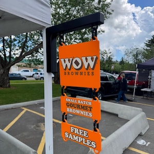 Puede incluir: Un letrero colgante vertical con tres paneles rectangulares naranjas conectados por eslabones negros. El panel superior dice WOW BROWNIES, el del medio SMALL BATCH GOURMET BROWNIES y el inferior dice FREE SAMPLES en letras blancas.