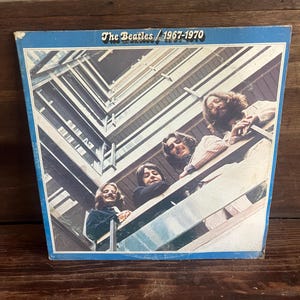 May include: A square vinyl record album cover titled The Beatles / 1967-1970. The cover features a low-angle photograph of four individuals looking down from a balcony with wooden railings, set against a blue border frame on a white background.