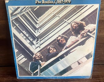 Könnte beinhalten: Ein quadratisches Vinyl-Schallplattencover mit dem Titel The Beatles / 1967-1970. Das Cover zeigt eine Froschperspektive von vier Personen, die von einem Balkon mit Holzgeländer herunterschauen, eingerahmt von einem blauen Rand auf weißem Grund.
