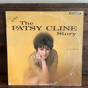 May include: A vintage vinyl record album cover titled The Patsy Cline Story, a deluxe 2-record set by MCA Records. The cover features a portrait of Patsy Cline against a yellow background, with handwritten text in the upper left corner.