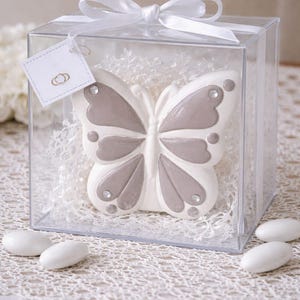 Può includere: Un oggetto decorativo a forma di farfalla bianca e grigia con dettagli in strass, presentato in una scatola quadrata trasparente. La scatola è legata con un nastro bianco e contiene imbottitura bianca. Un piccolo tag con un disegno di anelli dorati è appeso alla scatola.