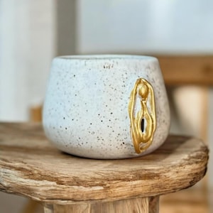 May include: A speckled white ceramic mug with a gold-colored vulva design on the side. The mug has a rounded shape and sits on a wooden surface. The gold design adds a decorative touch to the mug.