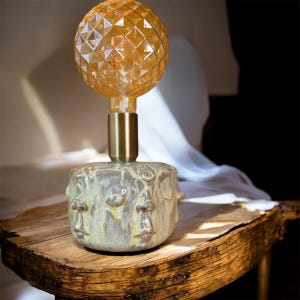 Peut inclure: Une lampe de table unique avec une ampoule sphérique ambrée texturée. L'ampoule est fixée sur une base de couleur laiton, elle-même montée sur un socle en céramique avec un motif de visage. La lampe est posée sur une surface en bois.