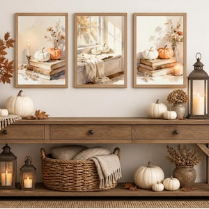 May include: A long wooden console table featuring three drawers, topped with three framed wall art prints of pumpkins and books. The display includes a vase of autumn leaves, white pumpkins, woven baskets with blankets, and metal lanterns with candles.