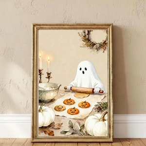 May include: A framed wall art print featuring a white ghost character rolling out dough on a table. The scene includes four pumpkin-shaped cookies, two white pumpkins, a mixing bowl, and two lit candles in brass holders, all set against a beige background.