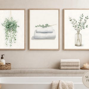 May include: A set of three framed watercolour prints with botanical themes. One print shows a hanging plant, another depicts folded towels with eucalyptus, and the third displays eucalyptus in a glass vase. The frames are light brown.