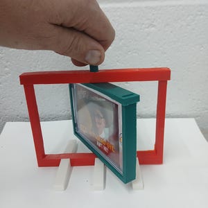 May include: A red and green 3D-printed display stand holding a framed image. The frame is green, and the stand is red. The image has the text "BAD EYE, AIN'T HE?" The stand is supported by white rectangular blocks.