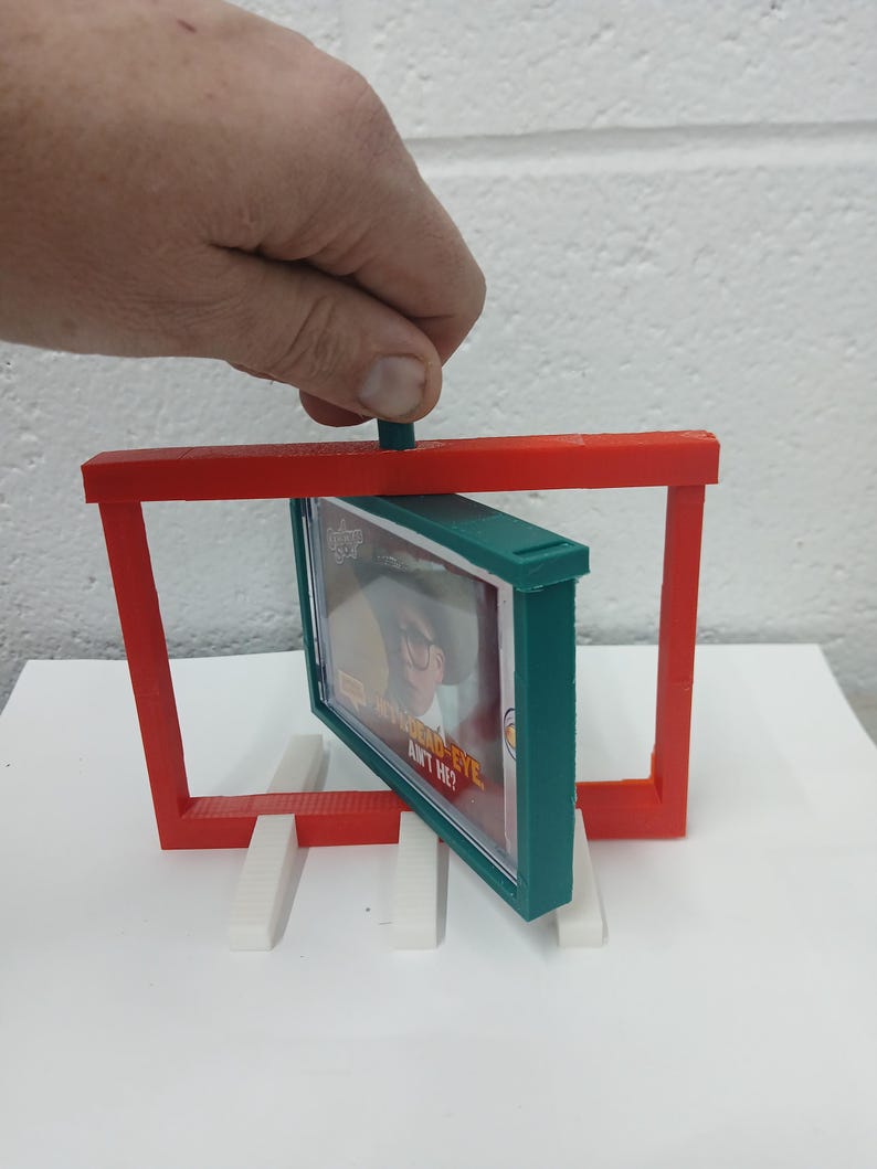 May include: A red and green 3D-printed display stand holding a framed image. The frame is green, and the stand is red. The image has the text "BAD EYE, AIN'T HE?" The stand is supported by white rectangular blocks.
