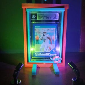 May include: A framed sports trading card featuring Jaylen Waddle in a Miami Dolphins uniform. The card is graded and encased in a clear plastic holder, displayed in a neon-lit orange and teal frame with two small spotlights.