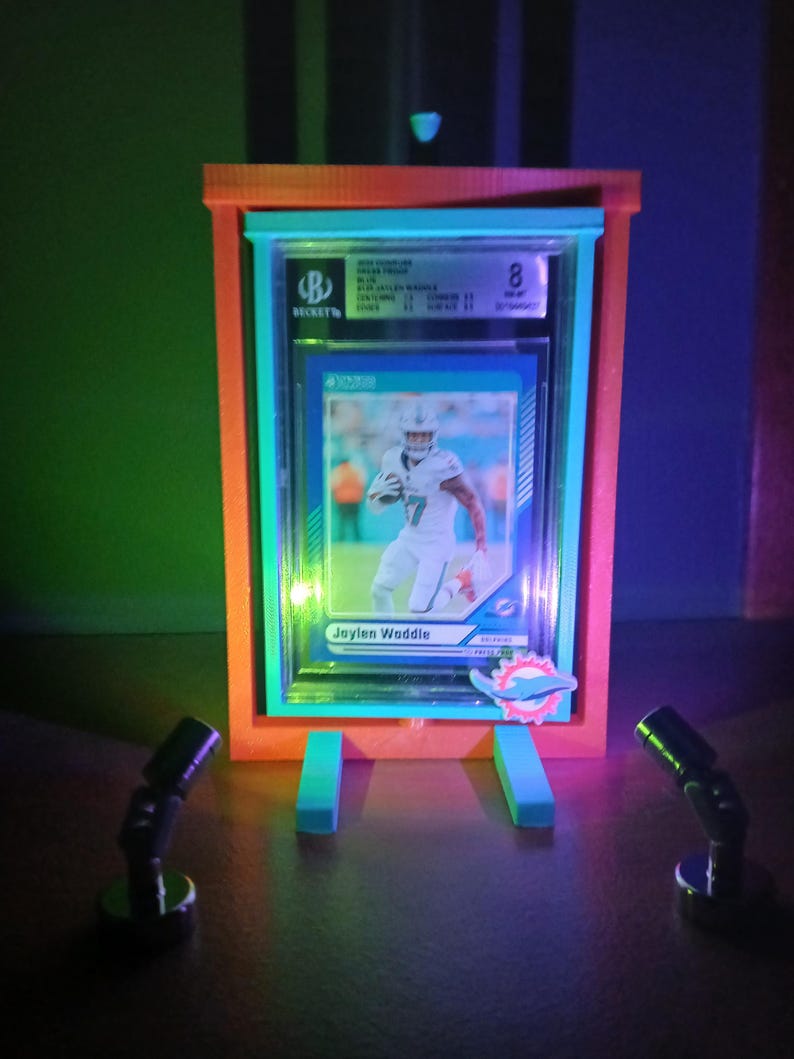 May include: A framed sports trading card featuring Jaylen Waddle in a Miami Dolphins uniform. The card is graded and encased in a clear plastic holder, displayed in a neon-lit orange and teal frame with two small spotlights.