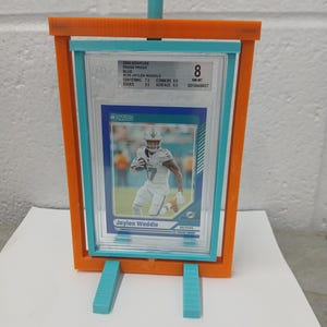 May include: An orange and teal display stand holding a graded Jaylen Waddle football card. The card is encased in a clear plastic holder. The stand has teal support legs and a teal top support.