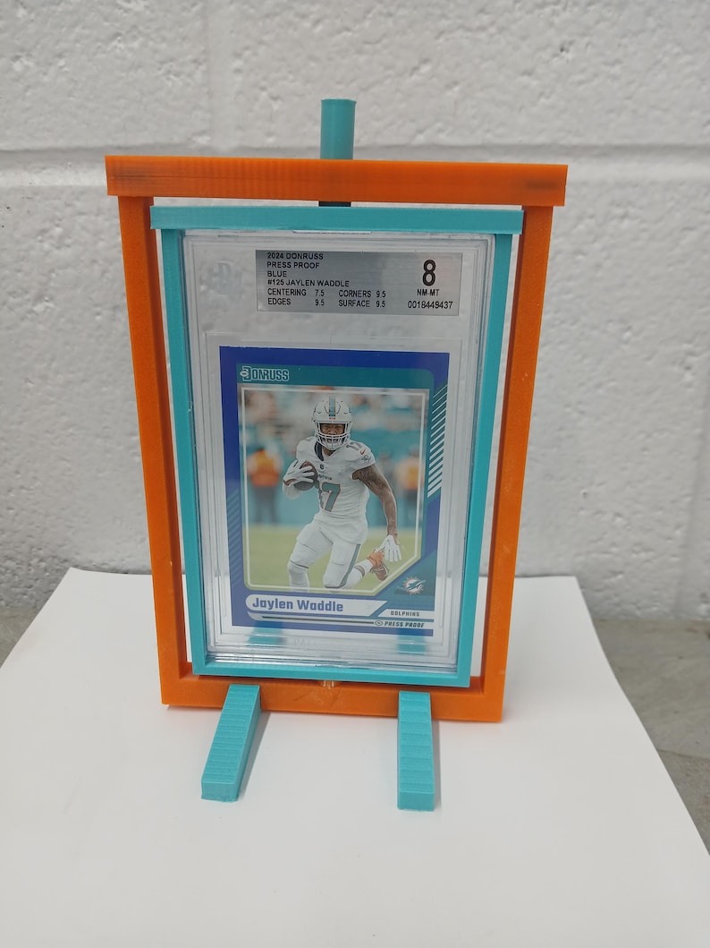 May include: An orange and teal display stand holding a graded Jaylen Waddle football card. The card is encased in a clear plastic holder. The stand has teal support legs and a teal top support.