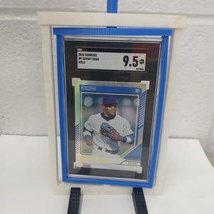 May include: A baseball card of Sammy Sosa, graded 9.5, displayed in a clear holder with a blue border. The card is set in a white and blue stand, with black support legs.