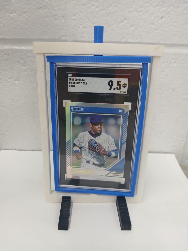 May include: A baseball card of Sammy Sosa, graded 9.5, displayed in a clear holder with a blue border. The card is set in a white and blue stand, with black support legs.