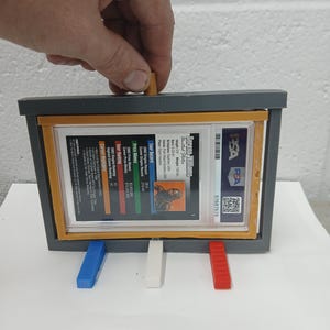May include: A gray display case with a clear window holding a Serena Williams trading card. The case has blue, white, and red supports. The card is graded by PSA and includes player stats. The case is being held by a hand.