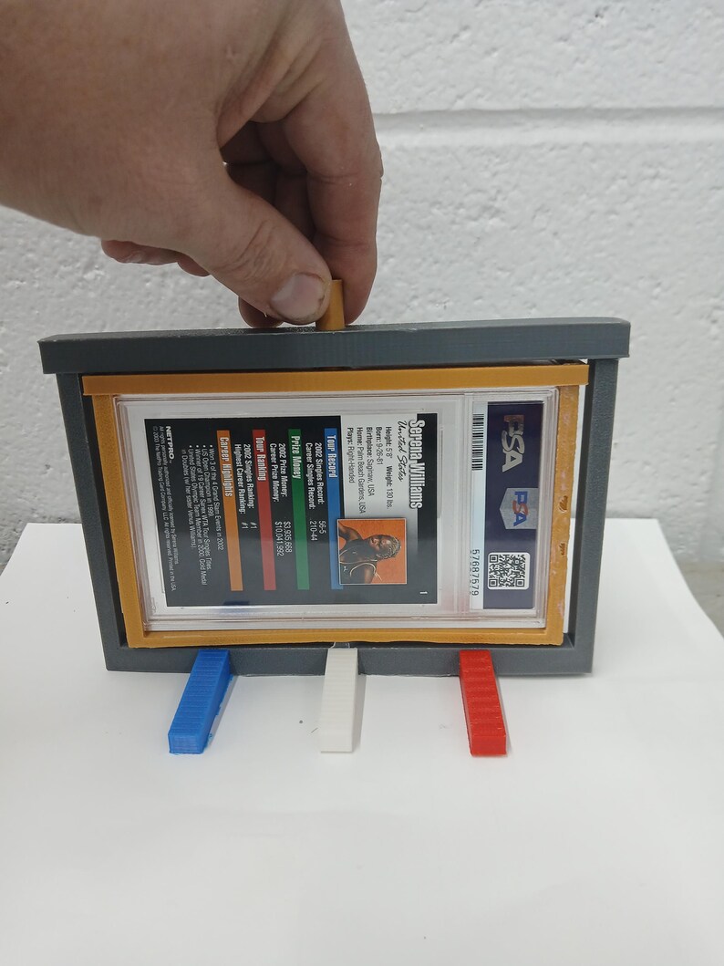 May include: A gray display case with a clear window holding a Serena Williams trading card. The case has blue, white, and red supports. The card is graded by PSA and includes player stats. The case is being held by a hand.