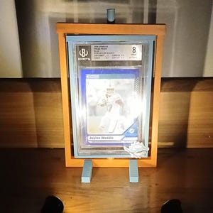 May include: A graded sports trading card of Jaylen Waddle, encased in a clear plastic holder. The card is displayed in an orange wooden frame with light blue supports. The Beckett grading label is visible.