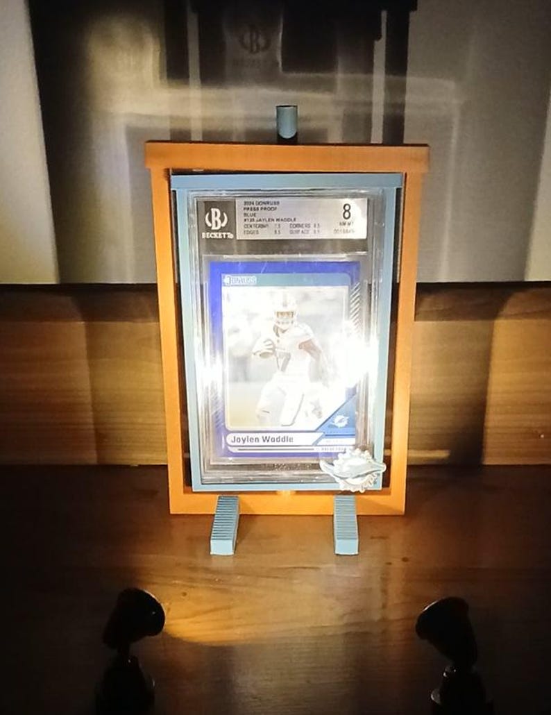 May include: A graded sports trading card of Jaylen Waddle, encased in a clear plastic holder. The card is displayed in an orange wooden frame with light blue supports. The Beckett grading label is visible.