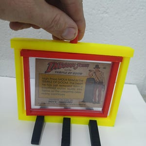 May include: A yellow and red 3D-printed frame holding an Indiana Jones trading card. The card features an illustration of Mola Ram from the Temple of Doom. The frame has three black supports.