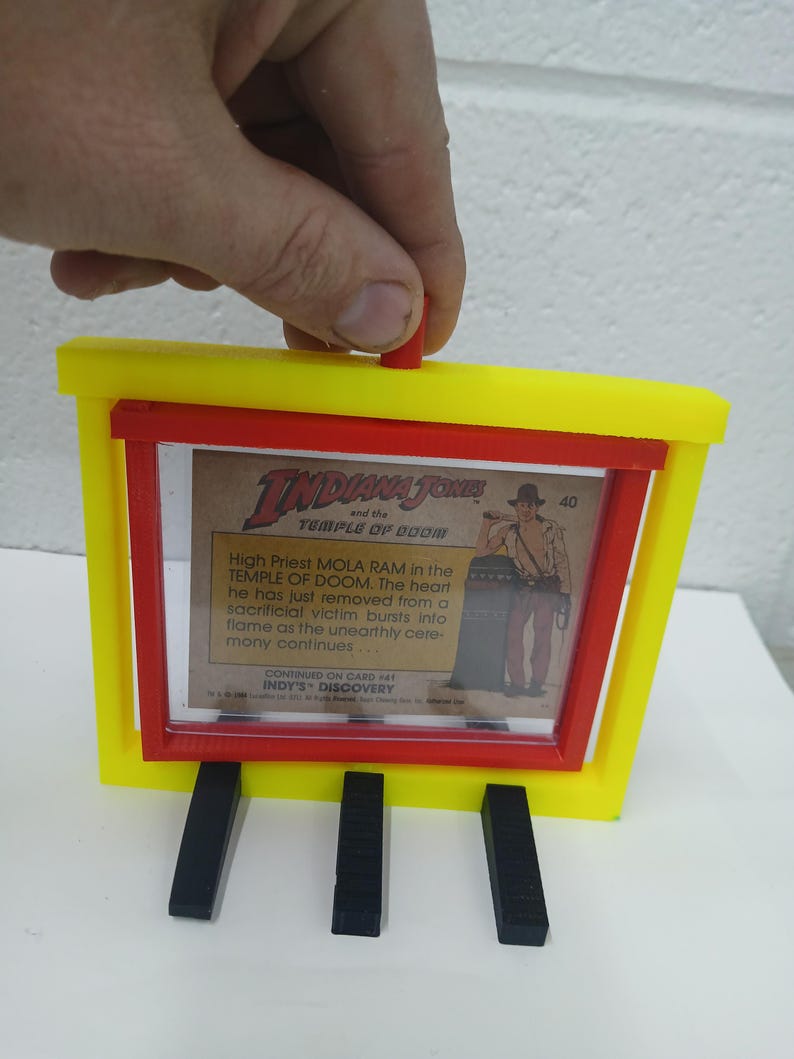 May include: A yellow and red 3D-printed frame holding an Indiana Jones trading card. The card features an illustration of Mola Ram from the Temple of Doom. The frame has three black supports.