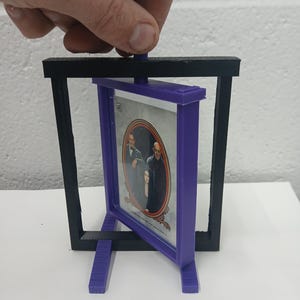 May include: A 3D-printed picture frame with a purple frame holding a picture of a family. The frame is held within a black rectangular stand. The frame is being held by a hand.