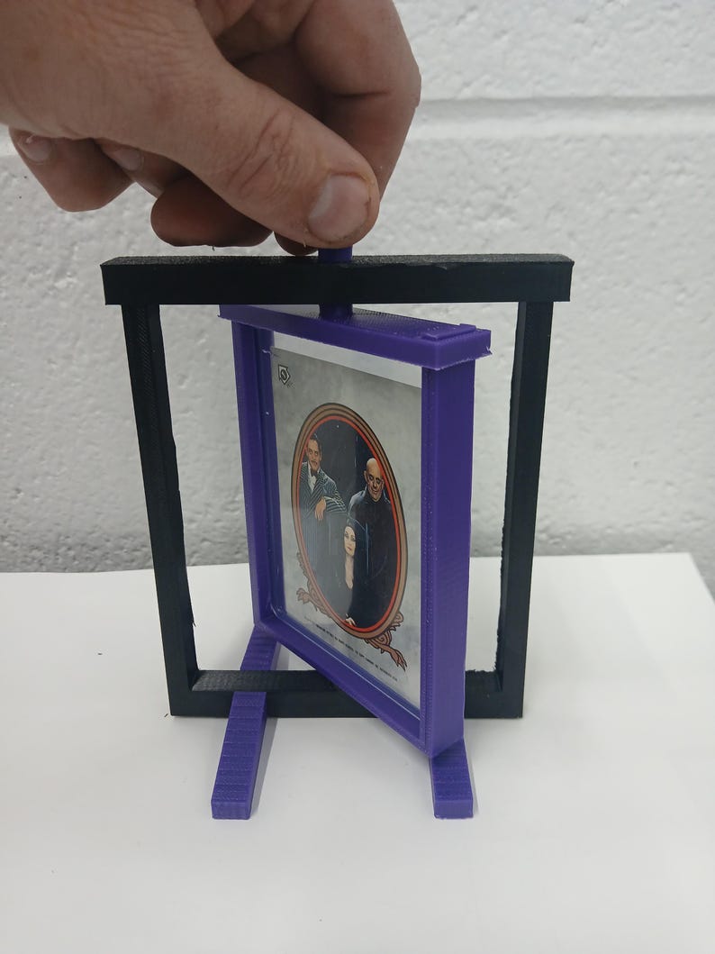 May include: A 3D-printed picture frame with a purple frame holding a picture of a family. The frame is held within a black rectangular stand. The frame is being held by a hand.
