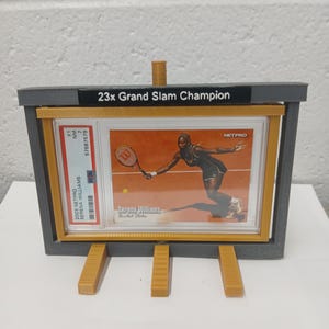 May include: A gray and yellow display stand holding a Serena Williams trading card. The card features Serena Williams playing tennis. The stand has the text "23x Grand Slam Champion" on top. The card is graded and encased.