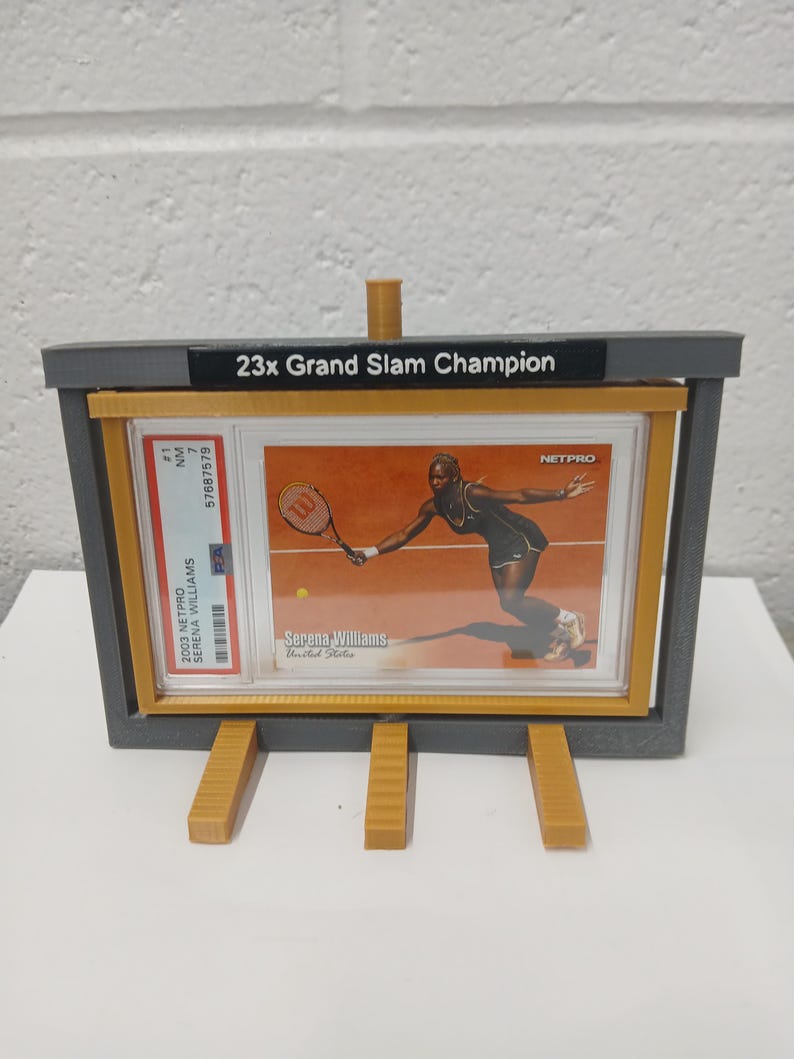 May include: A gray and yellow display stand holding a Serena Williams trading card. The card features Serena Williams playing tennis. The stand has the text "23x Grand Slam Champion" on top. The card is graded and encased.