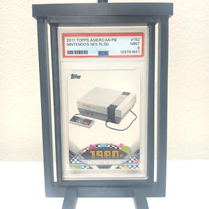 May include: A black display stand holding a graded trading card. The card features a Nintendo NES console and controller, with the text "2011 TOPPS AMERICAN PIE NINTENDO'S NES RLSD." and "1980s NES RELEASED". The card is graded as MINT 9.