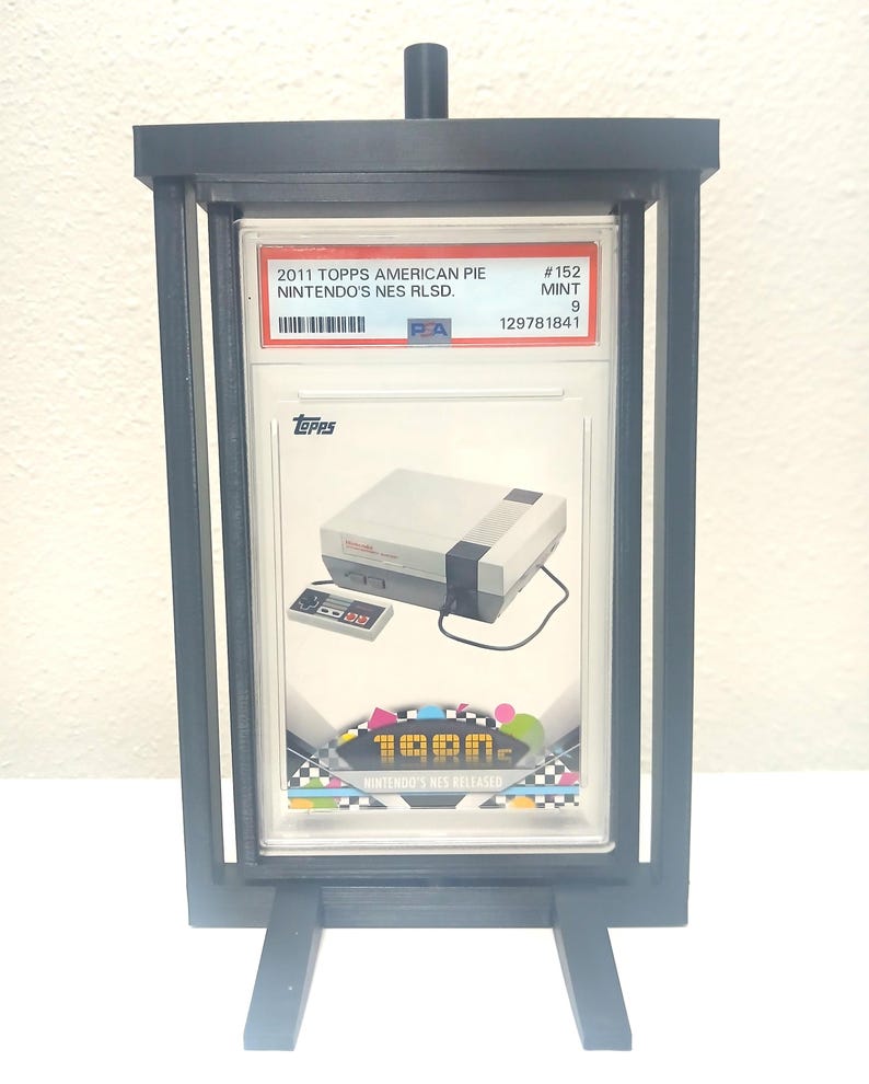 May include: A black display stand holding a graded trading card. The card features a Nintendo NES console and controller, with the text "2011 TOPPS AMERICAN PIE NINTENDO'S NES RLSD." and "1980s NES RELEASED". The card is graded as MINT 9.