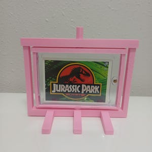 May include: A pink, 3D-printed easel-style frame holding a "Jurassic Park" graphic. The frame has a rectangular opening to display the graphic, with a small support at the top and three legs at the bottom. The graphic features a red dinosaur silhouette.