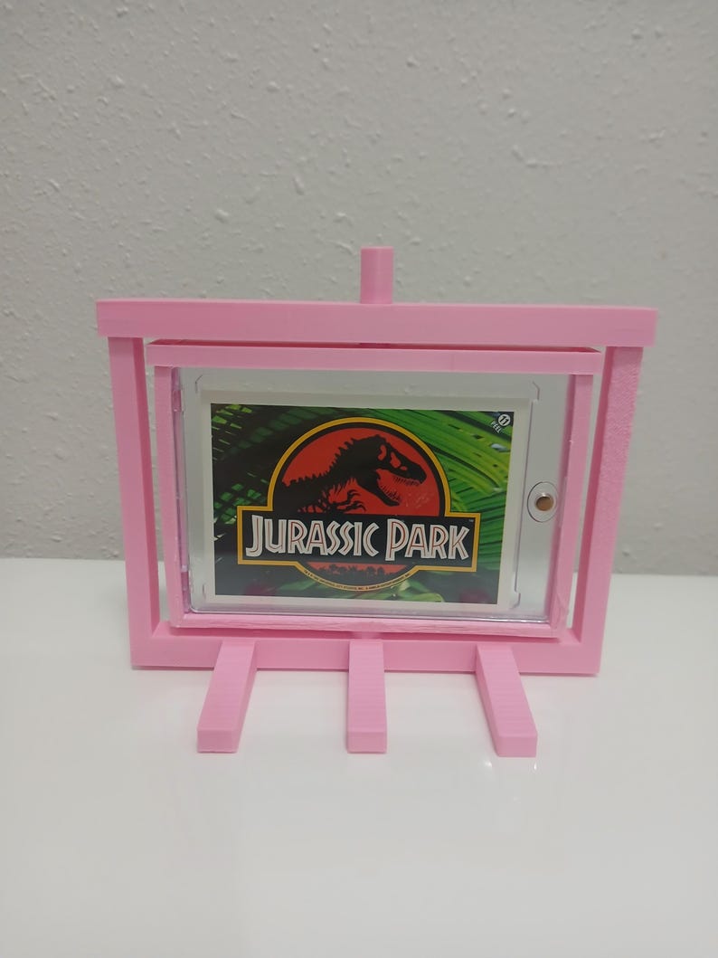 May include: A pink, 3D-printed easel-style frame holding a "Jurassic Park" graphic. The frame has a rectangular opening to display the graphic, with a small support at the top and three legs at the bottom. The graphic features a red dinosaur silhouette.