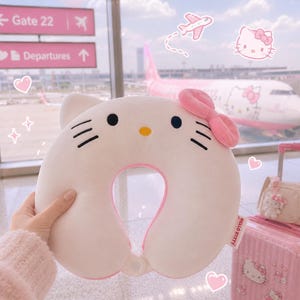 May include: A white U-shaped travel neck pillow featuring the Hello Kitty character face with black eyes, a yellow nose, black whiskers, and a pink bow. The pillow has a soft texture and a small white tag with the text Hello Kitty on the side.