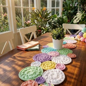Peut inclure: Une table en bois est ornée d'un chemin de table au crochet fait à la main. Le chemin de table présente diverses pièces de crochet circulaires aux couleurs pastel. Une petite plante en pot et des serviettes pliées sont également sur la table, dans une pièce ensoleillée avec des plantes.
