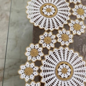 Puede incluir: Tapetes de ganchillo blancos y dorados dispuestos en un patrón floral. Los tapetes están hechos de hilo blanco con detalles dorados, creando un diseño delicado y elegante. Se colocan sobre una superficie de vidrio.