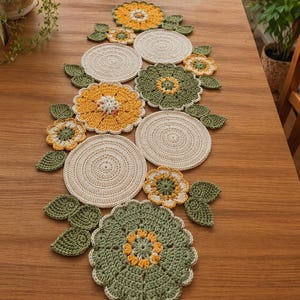 以下が含まれることがあります： 手作りのかぎ針編みのテーブルランナー。花柄のデザインで、黄色、緑、クリーム色の花と葉が含まれています。木製のテーブルの上に置かれています。