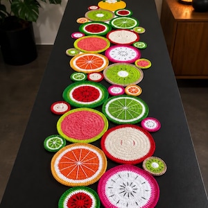 May include: A table runner composed of numerous circular crochet coasters featuring various fruit designs including watermelon, orange, lime, kiwi, and dragon fruit. The colorful, textured yarn pieces are arranged in a long, decorative cluster.