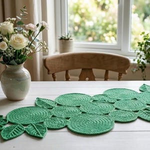Könnte beinhalten: Ein grüner gehäkelter Tischläufer mit einem Blatt- und Kreismuster. Der Läufer liegt auf einem weißen Tisch, im Hintergrund eine Vase mit weißen Rosen und Grün. Der Läufer ist etwa 152 cm lang.