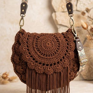 Peut inclure: Un sac bandoulière en crochet marron doté d'un rabat festonné et d'une longue frange. Le sac comprend une bandoulière réglable et amovible avec un motif géométrique multicolore et des attaches en similicuir marron foncé.