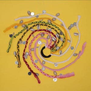 Puede incluir: Una disposición en espiral de coloridas trenzas para el cabello, adornadas con conchas marinas y monedas plateadas, sobre un fondo amarillo brillante. Las trenzas son de color rosa, rojo, verde, amarillo y blanco, con una goma negra en el centro.