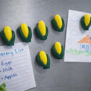Può includere: Una collezione di calamite a forma di mais con mais giallo e foglie verdi. È allegata una lista della spesa con "Lista della spesa" e articoli, insieme al disegno di un bambino di una casa. Le calamite sono su un frigorifero argentato.