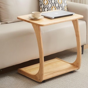 May include: A light-coloured wooden side table with a rectangular top and a lower shelf. A laptop and a teacup with saucer sit on the table. The table has curved legs and is positioned next to a beige sofa.