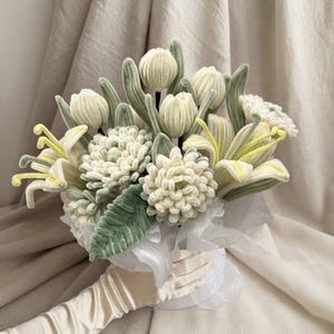 May include: A bouquet of handcrafted crochet flowers in shades of cream, pale yellow, and sage green. The arrangement includes rounded buds, textured pom-pom style blossoms, and long-petaled lilies with green leaves, all wrapped in white tissue paper.