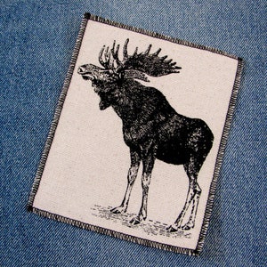 May include: A black and white illustration of a moose with large antlers on a white background. The moose is standing on a light brown surface. The illustration is surrounded by a black stitched border.