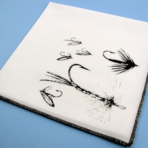 May include: A white cotton tea towel with a black and white illustration of fishing flies. The flies are arranged in a cluster with hooks and feathers.