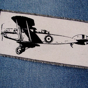 May include: A rectangular patch with a black and white biplane design. The biplane has a propeller, wings, and a roundel. The patch is edged with black stitching and is set against a denim background.