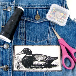 May include: A denim jacket with a black thread spool, a container of colorful pins, a pair of pink scissors, and a black and white duck patch.