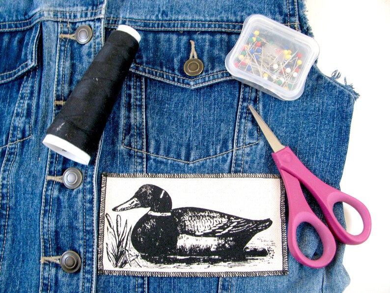 May include: A denim jacket with a black thread spool, a container of colorful pins, a pair of pink scissors, and a black and white duck patch.