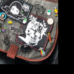 May include: A gray canvas bag with various pins and patches. The patches include a skull with flowers, a black and white illustration of a woman's face, and a patch that says "Put the dogs to sleep".
